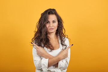 Holding hands crossed pointing index fingers aside, showing two directions, forefingers away. Young attractive woman, dressed white blouse, with brown eyes, curly hair, yellow background