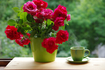 Obraz na płótnie Canvas A bouquet of red roses in a green vase and a cup of tea on the windowsill.