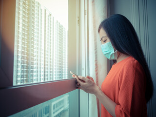 Asian woman, who is wearing a mask, is using her mobile phone by the room's window.
