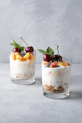 Healthy breakfast in a glass with fresh fruits: pomegranate, cherry, nectarines, honeysuckle, yogurt and granola on a grey background. Shallow depth of field with selective focus