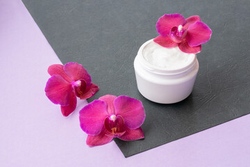 Obraz na płótnie Canvas White container with cream for face and body with three magenta colored orchid flowers on purple and black background. Concept of delicate or eco friendly cosmetics