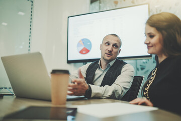 Business meeting in a boardroom with a selective focus on a caucasian man entrepreneur talking with his female colleague; a laptop, paper coffee cup, and a paper sheets on the table, LCD screen behind