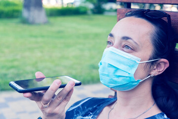 Young woman in protective mask holding a smartphone. Woman siting on bench in green park with...