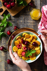 In the hands of a woman breakfast. Mini pancakes with cherries and honey on a rustic wooden table. Top view flat lay background.