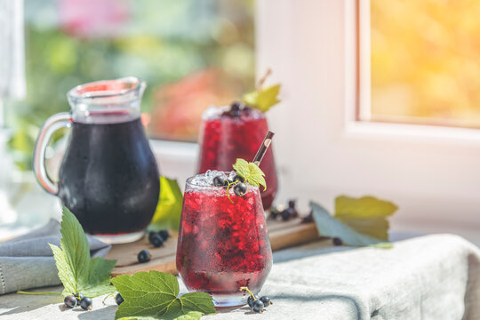 Two glass of cold ice black currant juice with ripe berries and green leaves on table in sunny room near window with garden outside