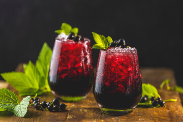 Two glass of cold ice black currant juice or cocktail with ripe berries and green leaves on dark...