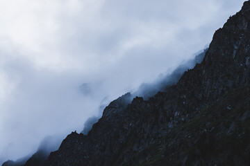 Dramatic bleak fog among giant rocky mountains. Ghostly atmospheric view to big cliff in cloudy sky. Low clouds and beautiful rockies. Minimalist scenery in mysterious place at early foggy morning.