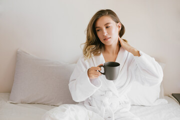 Obraz na płótnie Canvas Young beautiful woman wearing white bathrobe having breakfast in bed with coffee and croissant and fresh fruits in cozy bedroom.