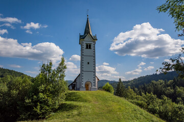 Small catholic church on hilltop. Bright sunny summer day. Grass covering landscape. In background hills covered with trees. Puffy clouds in the sky. Wide shot looking up