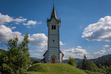Small catholic church on hilltop. Bright sunny summer day. Grass covering landscape. In background hills covered with trees. Puffy clouds in the sky. Wide shot looking up