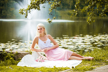 Portrait of a young blonde woman resting near a pond in a summer park