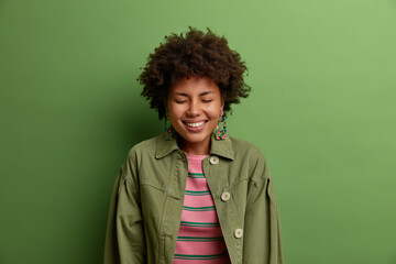 Fototapeta na wymiar Lovely positive young African American woman smiles broadly, shows white teeth, closes eyes, expresses happiness, enjoys nice talk, laughs at something funny, wears green jacket, stands indoor