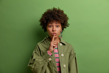 Mysterious beautiful woman with Afro hair presses index finger to folded lips makes hush gesture and asks to be quiet asks politely to keep silence prohibits talking during meeting wears green clothes - obrazy, fototapety, plakaty