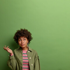Vertical shot of pensive dark skinned woman concentrated above, gestures with palm, thinks about creative solution, wears stylish clothes, isolated on green background, blank space for your promo