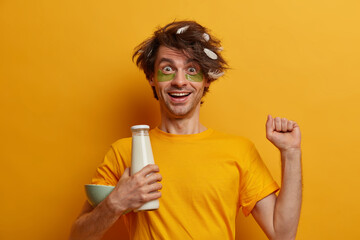 Happy positive man has morning snack, stretches and feels refreshed, wears collagen patches under eyes, has enough amount of sleep, messy hairstyle, poses over yellow wall. Breakfast tradition