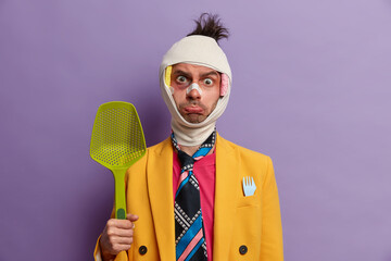 Bruised displeased man ready for fight and defence himself, poses with kitchenware, injured after...
