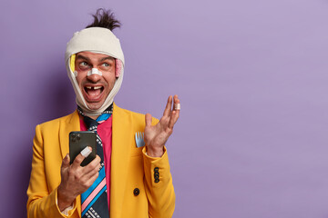 Photo of funny man with missing teeth, has bruises under eyes, beaten during corporative party, sort out relationships with someone, uses mobile phone for surfing net, isolated on purple background