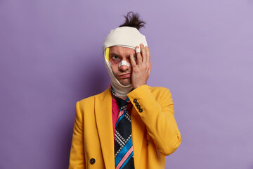 Sullen unhappy adult man hides his bruised face after got into car aacident, beaten by robbers, wears bandage around injured head, isolated over purple background. Male has brain concussion.