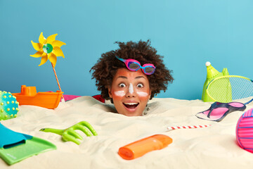 Happy surprised Afro American woman spends summer vacation at resort place on island, poses at sandy beach, sticks out head from sand, enjoys nice relaxation, got sunburn, uses sunblock cream.