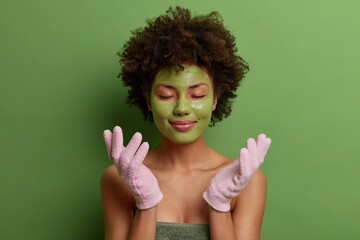 Calm relaxed beautiful woman applies avocado mask on face, wears bath gloves, closes eyes and waits for nice effect, forgets about all troubles with beauty procedures, has curly hair isolated on green