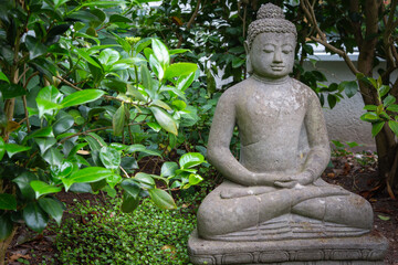 Full view Meditative Buddha stone sculpture in the lotus seat in a garden in front of green bushes