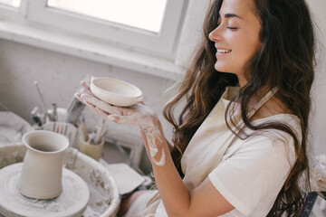 Young beautiful woman with long curly hair in white apron creating handmade ceramic bowl in a...