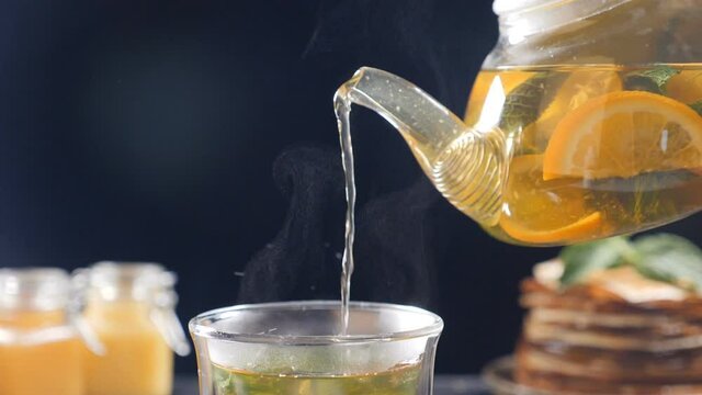 Pouring tea with orange, lemon and lime out of transparent tea pot into glass with nint. Tea set, glass teapot and cups. Brewing tea with honey and pancakes in background. Slow motion tea. Full hd