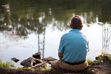 An elderly Caucasian man sitting on a riverbank on a trunk of a fallen tree and looking at the reflections in the water. Background with blurry background.