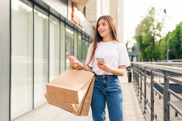 Young woman doing online shopping through mobile phone. Girl in fashion look holding bags near shoulder, looking at phone and smiling walking in the street