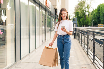 Young woman doing online shopping through mobile phone. Girl in fashion look holding bags near shoulder, looking at phone and smiling walking in the street