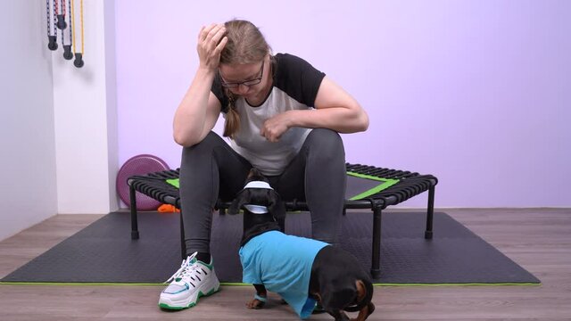 Very tired young Caucasian woman sits on the edge of little home trampoline and breathing heavily under the jumping. Funny black and tan dachshund comes up to her and they look at each other. Indoors.