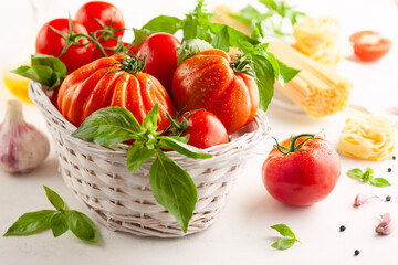 Fototapeta na wymiar Assorted tomatoes with basil in basket, garlic, spice and raw pasta for italian cuisine. Healthy food concept on white background.
