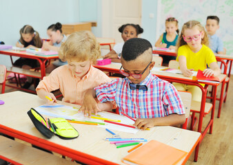 Fototapeta premium two small children of elementary school students are preparing for a lesson sitting at a Desk with colored pencils in their hands against the background of a group of children in the classroom.