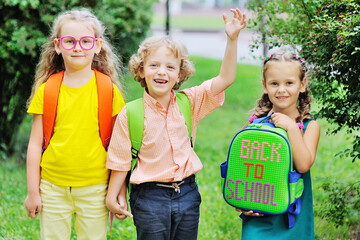 three school children-two girls and a curly-haired boy with school colored bags smiling hold hands and go to school. Back to school concept, September 1, knowledge day, education.