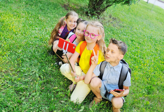 a group of school children sitting on the grass in colorful clothes and carrying school bags smile play and take photos on a smartphone camera on a selfie stick. Back to school