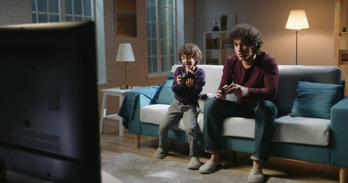 Funny asian siblings with curly hair are having fun at home, playing video games in front of tv, authentic father and son enjoying their hobby 4k footage