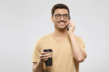 Young handsome businessman in beige t-shirt and eyeglasses, talking on phone with takeaway coffee cup in hands, isolated on gray background