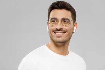 Young handsome man with short dark hair, wearing white t-shirt, eyeglasses and wireless earphones, looking aside with dreamful smile, isolated on gray background