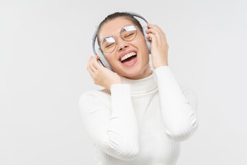 Fototapeta premium Happy excited girl wearing glasses and wireless headphones, laughing, listening to music, feeling positive and relaxed, isolated on gray background
