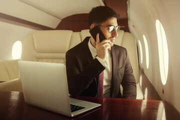 Rich man travelling first class in private jet, sitting in leather armchair at table, talking on phone with partners and looking through window