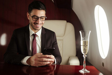 Rich businessman sitting on board of private jet in armchair at table, drinking champagne and сhecking business news on his phone