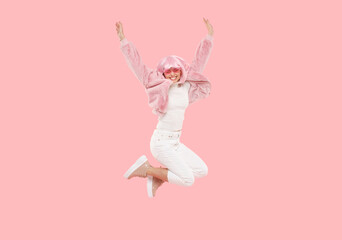 Young happy teenage girl wearing colored funky wig jumping in air, bending knees and lifting hands up, isolated on pink background