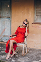 Stylish and elegant woman in a red dress is resting and smile near the house. sitting on a chair.