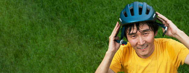 Banner with joyful asian man in yellow t-short holding a sport helmet over the head. Portrait against green grass. Solo travell, road trip and hyper-local travel concept.