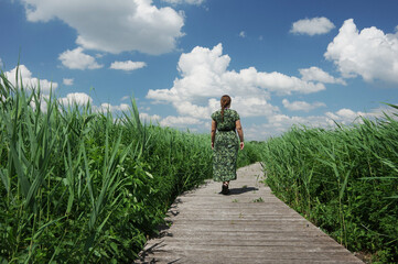 A girl in green dress walking on the  wooden path in the high green grass; blue sky, sunny day.