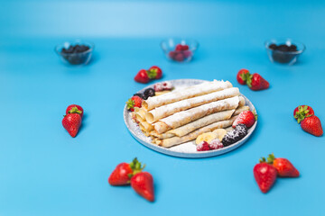 Close-up of a beautifully decorated serving plate full with French Crepes with strawberries around the plate and series inside. Homemade French crepes on blue background with free space for decoration