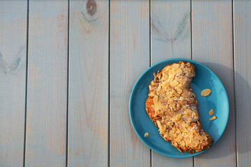 Tasty and sweet almond croissant laying on blue plate standing on light-blue wooden table in the day light with place for text.