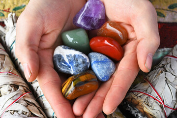 A top view image of a hand holding several chakra crystals with white sage.