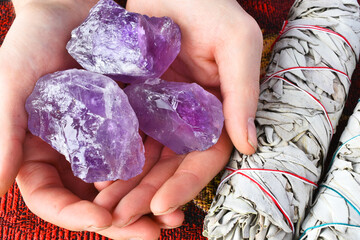 A close up image of a hand holding three purple amethyst crystals next to two white sage bundles. 