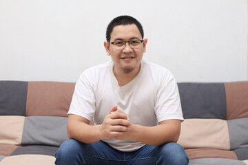 Handsome young Asian man sitting at home on sofa, smiling happy, looking at camera